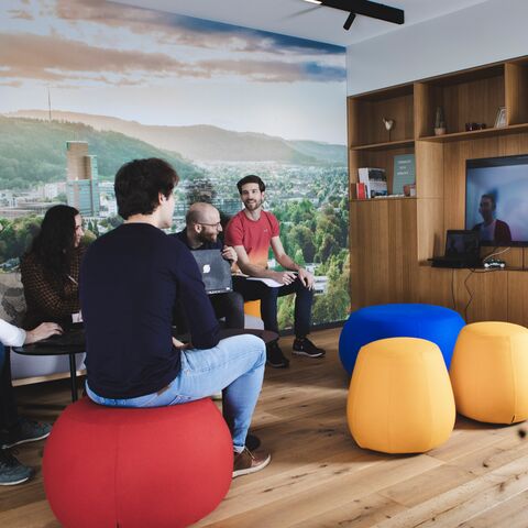 Eine Gruppe von fünf Menschen sitzt auf bunten Hockern in einem modernen Raum und betrachtet einen Videocall auf einem grossen Bildschirm.