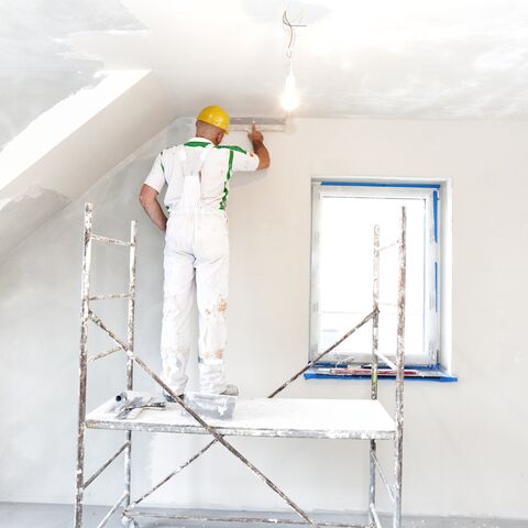 Un imbianchino con un casco giallo e una tuta bianca è in piedi su una scala e dipinge un soffitto bianco quando la luce è accesa.