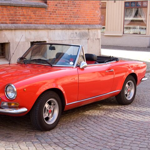 Cabriolet rossa d'epoca parcheggiata su una strada acciottolata davanti a un edificio in mattoni con finestre e porte in legno.