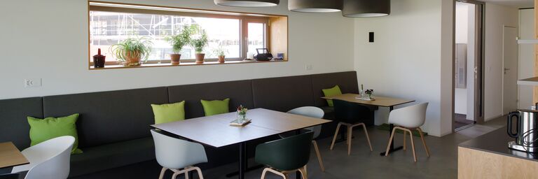 Un salone moderno con grandi finestre, piante, panchine grigie, cuscini verdi e tavoli in legno con sedie bianche e nere.