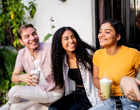 Three smiling friends are sitting outside on a bench enjoying drinks; one is holding coffee, the others smoothies, surrounded by plants.