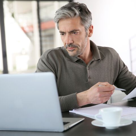 Mann am Schreibtisch schaut ernst in seinen Laptop und hält Dokumente und einen Stift in der Hand.