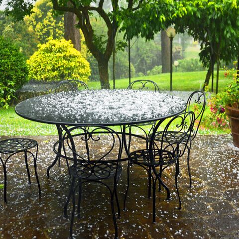 Un tavolo rotondo e quattro sedie in metallo nero su una terrazza coperta di chicchi di grandine, circondata da piante e alberi verdi.