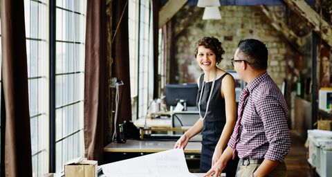A smiling woman talks to a colleague at her desk in an office.
