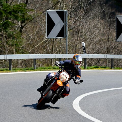 Al volante di una moto arancione, un motociclista in abiti neri e arancioni sta percorrendo in modo dinamico una curva su una strada vuota.