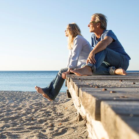 Ein Mann und eine Frau sitzen entspannt auf einem Holzsteg am Strand und blicken lächelnd aufs Meer unter klarem, blauem Himmel.