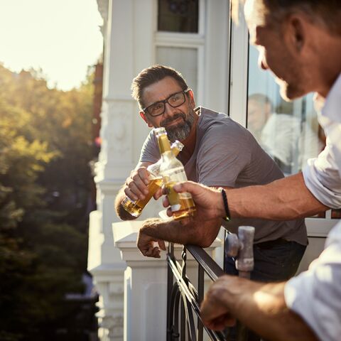 Due uomini sorseggiano bottiglie di birra su un balcone assolato mentre chiacchierano. Sullo sfondo si vedono alberi e case.