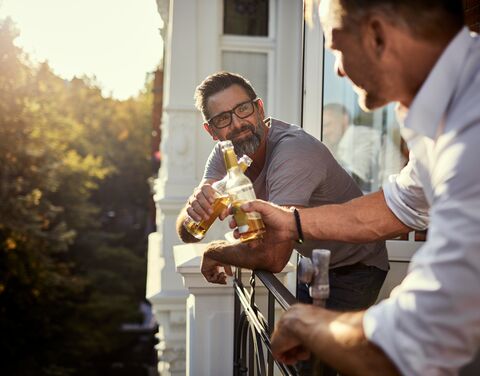 Zwei Männer stossen mit Bierflaschen auf einem sonnigen Balkon an, während sie sich unterhalten. Im Hintergrund sind Bäume und Häuser zu sehen.