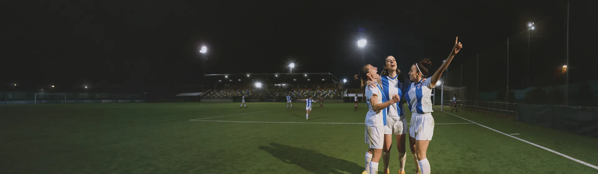 Trois joueuses de football exultent sur le terrain sous les projecteurs après avoir marqué un but.