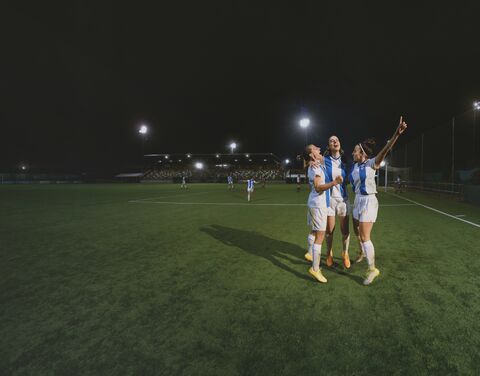 Three footballers cheer on the pitch under floodlights after scoring a goal.