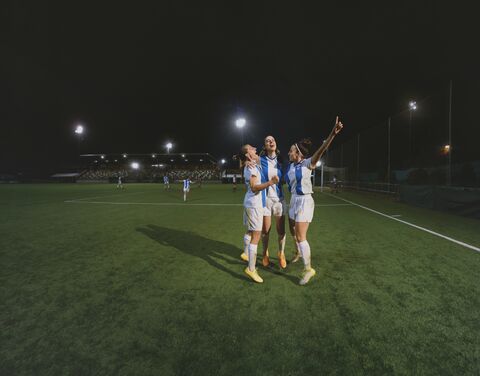 Three footballers cheer on the pitch under floodlights after scoring a goal.