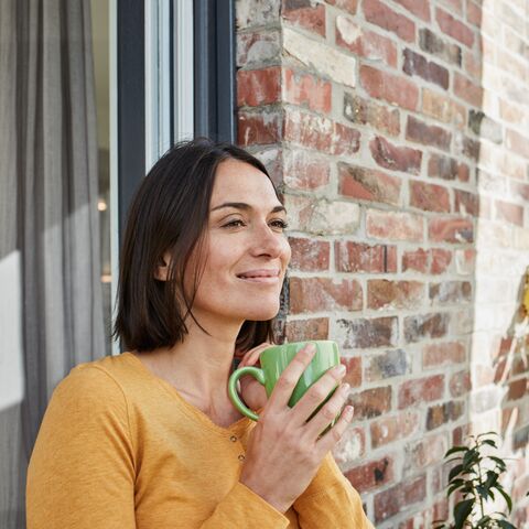 Junge Frau lehnt sich draussen am Fenster ihres Backsteinhauses an. Sie hält eine grüne Tasse in der Hand und lächelt.