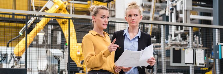 Zwei Frauen besprechen einen Plan in der Fabrikhalle mit einem Industrieroboter