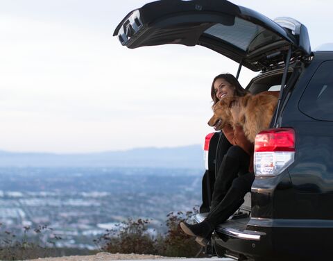 A woman sits with a dog in the open trunk of a car and enjoys the view of a vast landscape in the background.