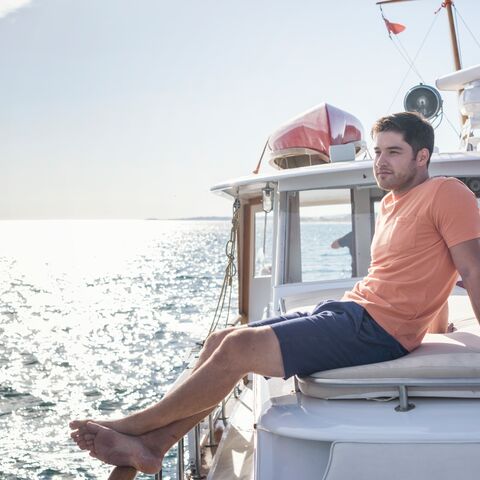 A young man in a red shirt sits contentedly on the roof of his motor yacht, looking out to the sea.