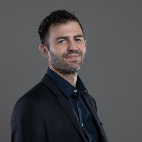 Un simpatico giovane con una barba ben curata sorride alla telecamera. Indossa una giacca color antracite e sotto una camicia blu scuro.