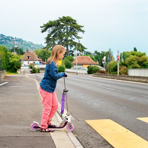 Ein Mädchen in blauer Jacke und rosa Hose steht mit einem lilafarbenen Roller an einem Zebrastreifen auf einer leeren Strasse.