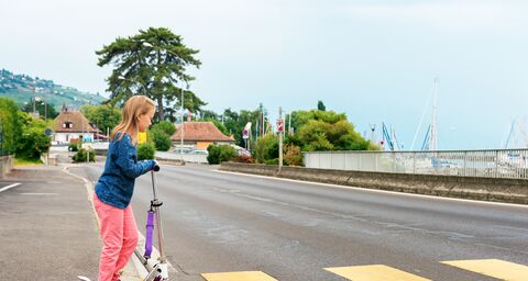 Ein Mädchen in blauer Jacke und rosa Hose steht mit einem lilafarbenen Roller an einem Zebrastreifen auf einer leeren Strasse.