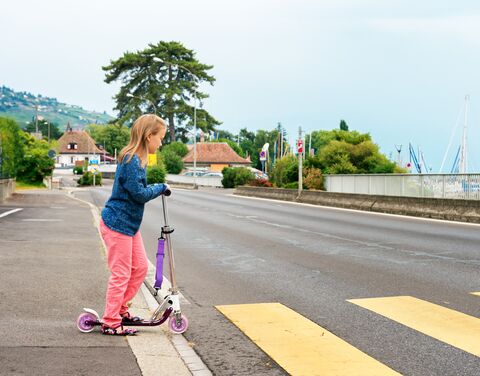 Una ragazza in giacca blu e pantaloni rosa sosta sulle strisce pedonali su una strada vuota con uno scooter viola.