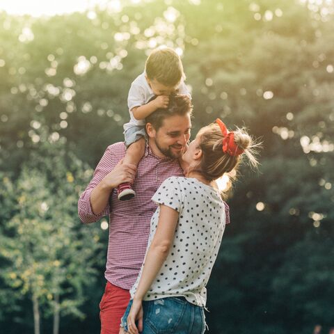 A smiling couple is standing outside in front of some trees. The man has a child sitting on his shoulders while his wife gives him a kiss.