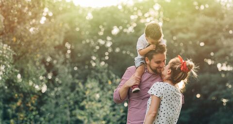 A smiling couple stands outside in front of trees, the man carrying a child on his shoulders while the woman gives him a kiss.