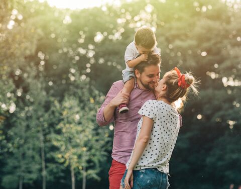 Un couple souriant se tient devant des arbres, l’homme porte un enfant sur ses épaules tandis que la femme l’embrasse.