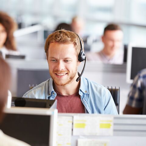 Ein junger Mann mit Headset lächelt und sitzt in einem modernen Büro, während er mit einer Person vor ihm kommuniziert.