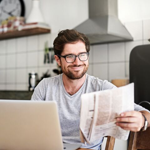 Giovane uomo con gli occhiali è seduto in cucina davanti al suo laptop e legge il giornale.