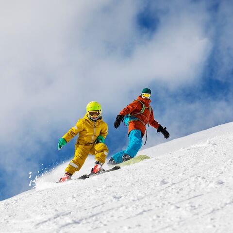 Un uomo in pantaloni da sci blu e giacca arancione fa snowboard dietro a un bambino in tuta gialla sugli sci. Alle loro spalle, il cielo è azzurro con qualche nuvola.