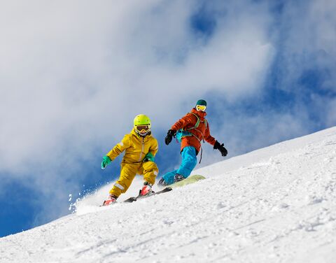 A man in blue ski pants and an orange jacket is snowboarding behind a child in a yellow ski suit on skis. Behind them, the sky is blue with a few clouds.