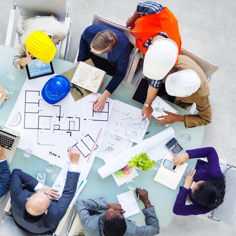 Un groupe de personnes portant des casques de chantier est assis autour d'une table et discute des plans de construction tout en prenant des notes et en utilisant des ordinateurs portables.