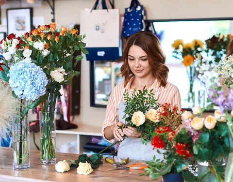 Une fleuriste avec un chemisier rayé dispose différents bouquets de fleurs sur une table en bois dans un magasin lumineux.