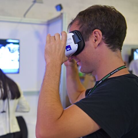 Ein junger Mann mit einem schwarzen T-Shirt setzt sich eine VR-Brille der AXA auf. Im Hintergrund ist unscharf eine Frau mit langen dunklen Haaren und einem weissen Blazer von hinten zusehen, die durch eine VR-Brille schaut und in der linken Hand einen schwarzen Joystick hat.