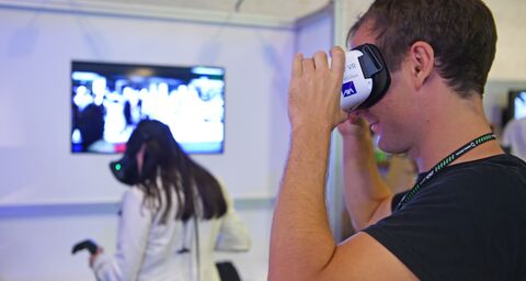 Ein junger Mann mit einem schwarzen T-Shirt setzt sich eine VR-Brille der AXA auf. Im Hintergrund ist unscharf eine Frau mit langen dunklen Haaren und einem weissen Blazer von hinten zusehen, die durch eine VR-Brille schaut und in der linken Hand einen schwarzen Joystick hat.