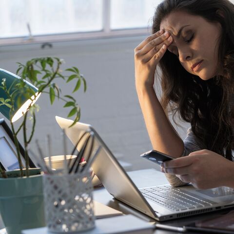 Eine Frau sitzt am Tisch vor dem Laptop und hat Ihr Handy in der Hand. Sie hält sich die Stirn und sieht erschöpft aus. 