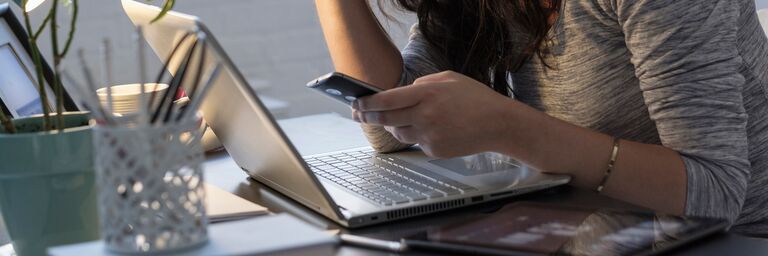 Eine Frau sitzt am Tisch vor dem Laptop und hat Ihr Handy in der Hand. Sie hält sich die Stirn und sieht erschöpft aus. 
