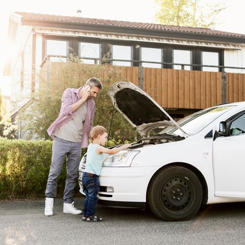 Un uomo telefona vicino a un’auto parcheggiata con il cofano aperto, mentre un bambino guarda curioso nel vano motore.