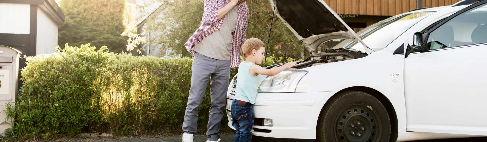 Un homme téléphone à côté d’une voiture parquée dont le capot est ouvert, tandis qu’un enfant regarde le compartiment moteur avec curiosité.