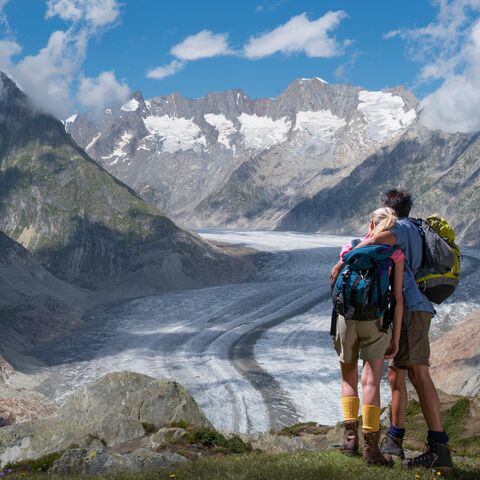 Zwei Wanderer stehen auf einem Felsvorsprung und blicken auf einen weiten Gletscher in einer malerischen Berglandschaft unter blauem Himmel.