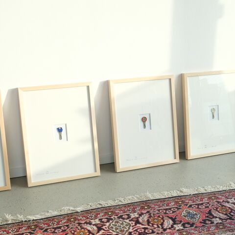 Fünf Bilderrahmen mit jeweils einem Schlüssel stehen auf dem Boden vor einer weissen Wand. Im Vordergrund liegt ein rotgemusterter Teppich.