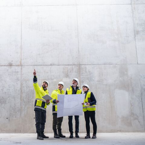 Quatre ingénieurs en bâtiment en tenue de protection et casqués discutent les yeux bleus dans un grand hall en béton, l'un d'eux pointe vers le haut.