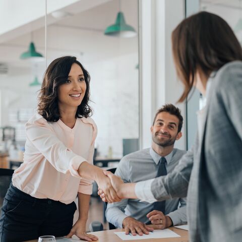 Tre persone in un ufficio moderno, una donna stringe la mano a un’altra donna, mentre un uomo guarda sorridendo.