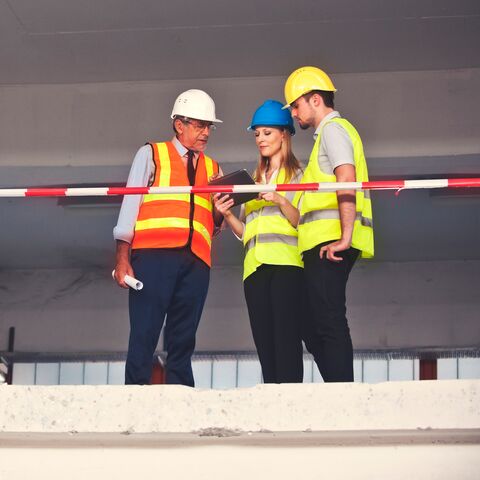Tre operai edili in abbigliamento protettivo discutono dei progetti in un cantiere. Indossano casco e giubbino di sicurezza e si trovano dietro una transenne.