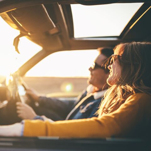 A smiling woman with sunglasses sits in her car with her hair blowing in the breeze while the evening sun streams in through the window.