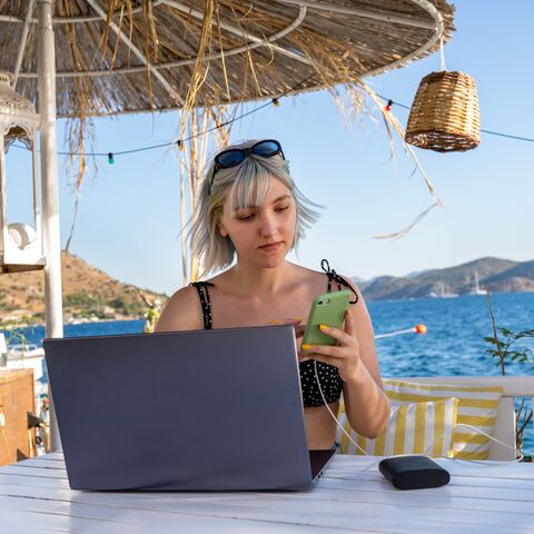 A woman sits under a thatched roof by the sea, working on a laptop and looking at her smartphone; mountains and boats in the background.