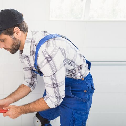 A plumber with a baseball cap and plaid shirt uses a wrench to fix a pipe in a bright room.