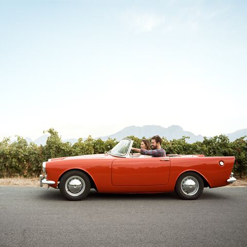 Une décapotable rouge roule sur une route de campagne le long d'un vignoble avec deux personnes qui profitent de la journée ensoleillée.