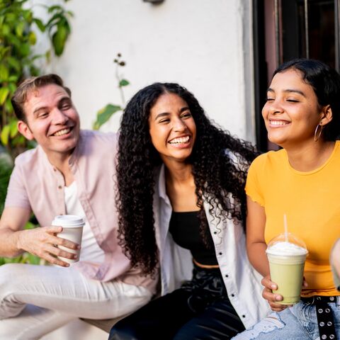 Drei lachende Freunde sitzen draussen auf einer Bank und geniessen Getränke; einer hält Kaffee, die anderen Smoothies, umgeben von Pflanzen.