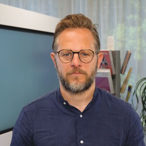 Ein Mann mit Brille, Bart und blauem Hemd steht in einem Eingangsbereich eines Unternehmens.