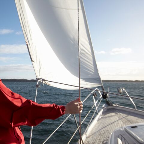 Una donna sorridente con fluenti capelli rossi si trova a bordo di una barca a vela, tenendo una corda e guardando il mare soleggiato.